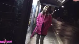 Blonde Frau im pinken Mantel steht nachts allein an einer beleuchteten Bushaltestelle